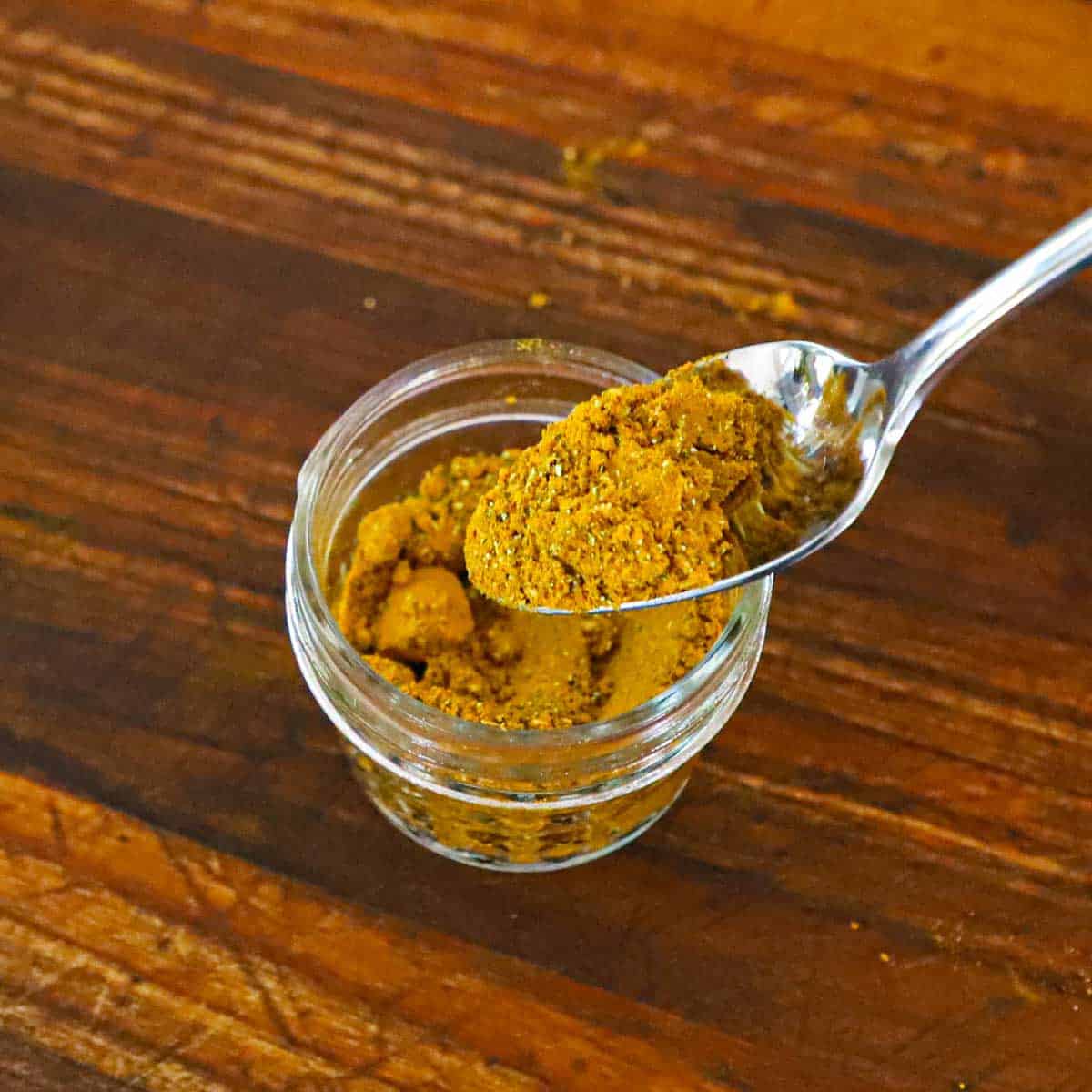 A person using a spoon to transfer homemade curry powder into a small glass bowl on a wooden cutting board.