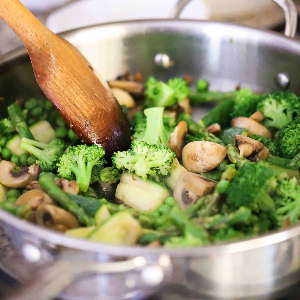 A person using a wooden spoon to stir cooked broccoli, asparagus, zucchini, and mushrooms in a large stainless steel skillet on a gas stove.