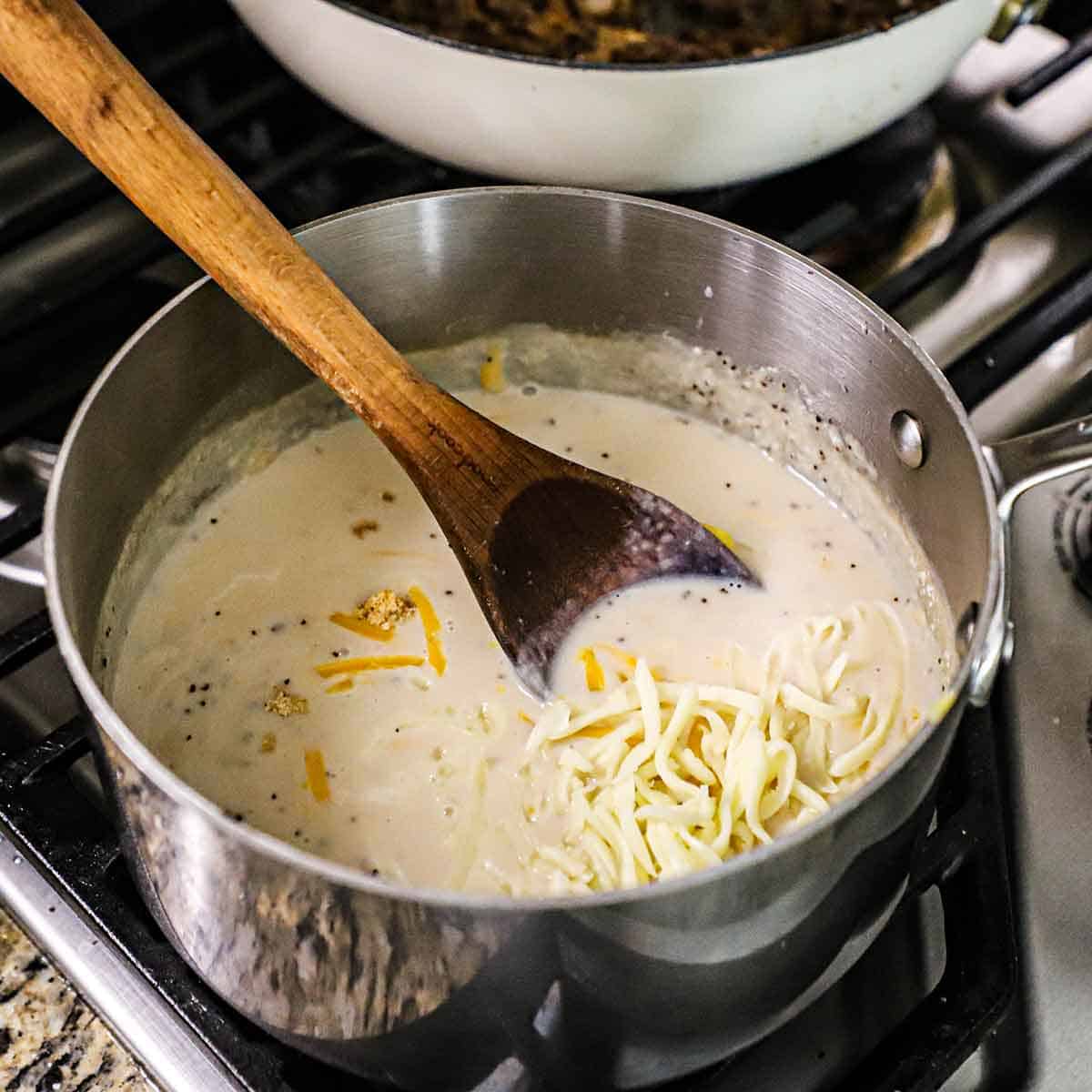 A silver saucepan filled with a bechamel sauce and shredded cheese being stirred with a large wooden spoon.