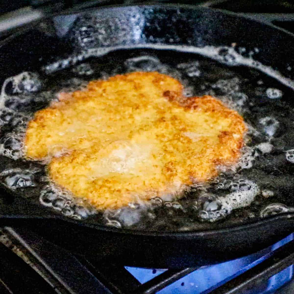 A straight-on view of a breaded pork tenderloin being fried in a large cast-iron skillet in hot oil on a gas stove.