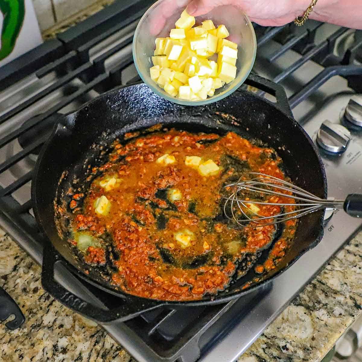 A person transferring cubed cold butter from a small glass bowl into a large cast-iron skillet on a gas stove filled with a simmering spiced garlic butter sauce.