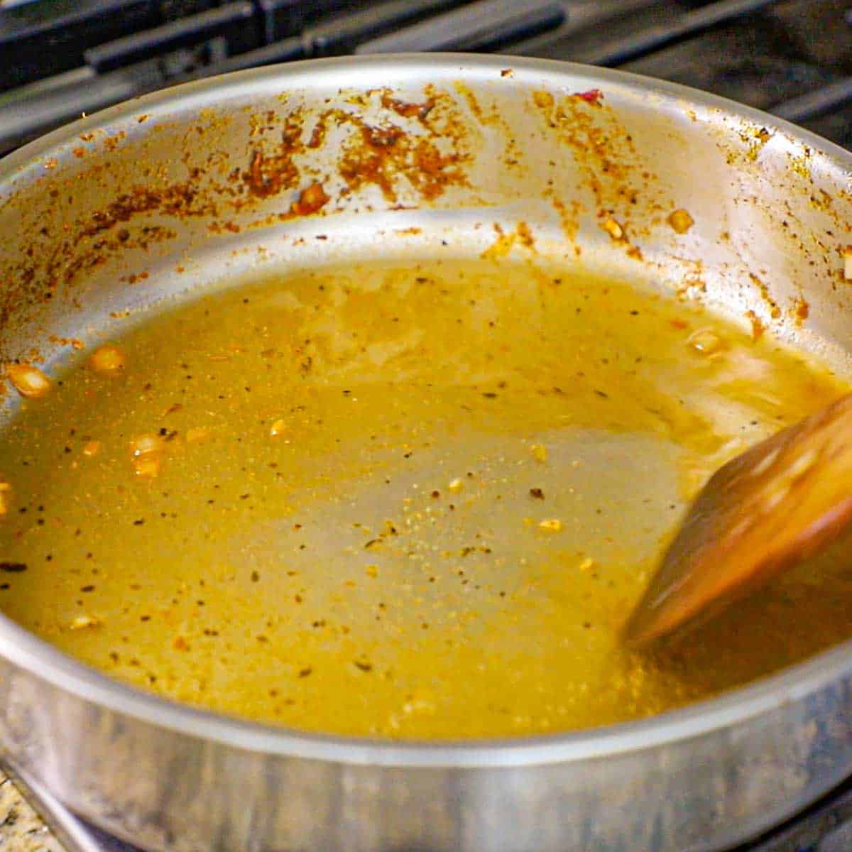 A person using a wooden spatula to deglaze a large stainless steel skillet that is filled with a thin layer of simmering white wine.