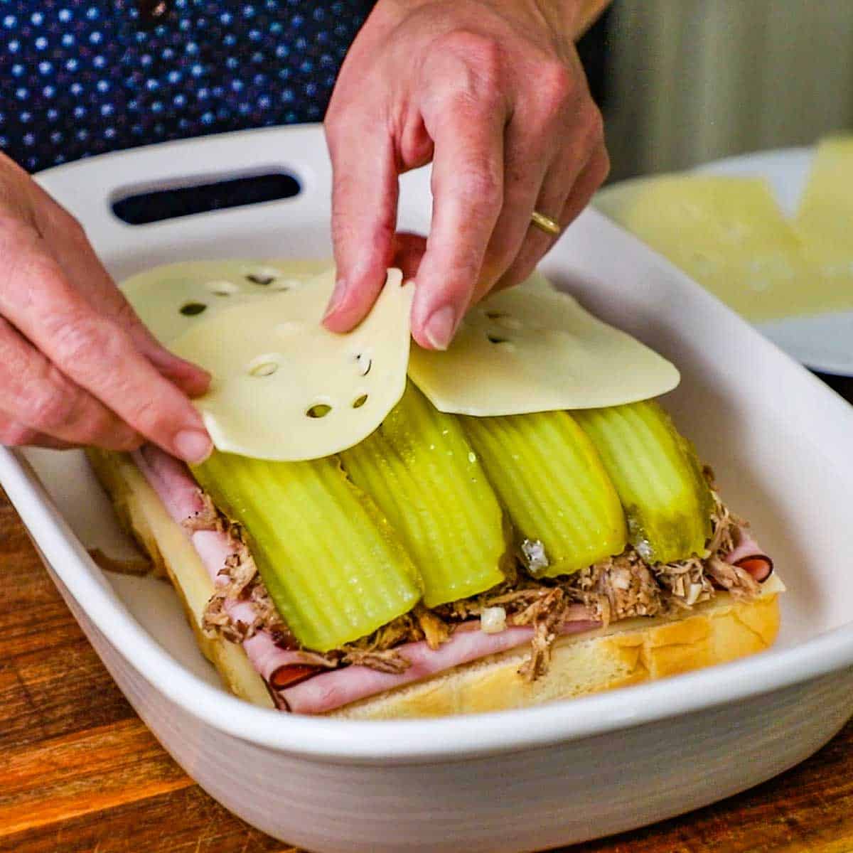 A person placing Swiss cheese slices over layers of ham, pork, and dill pickles on a layer of Hawaiian slider buns all in 9 by 13 inch baking dish.