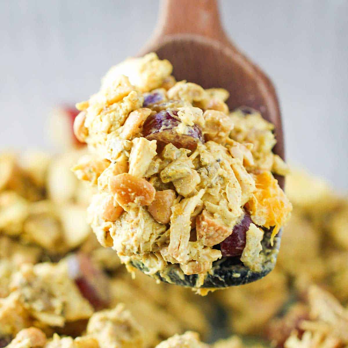 A person holding a large wooden spoon to lift a mound of curried chicken salad over a glass bowl filled with the same.