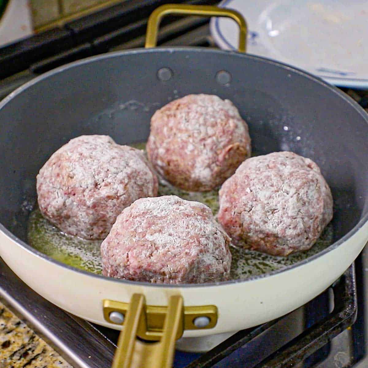 A straight-on view of four large Dutch meatballs simmering in a large non-stick skillet in melted butter on a gas stove.