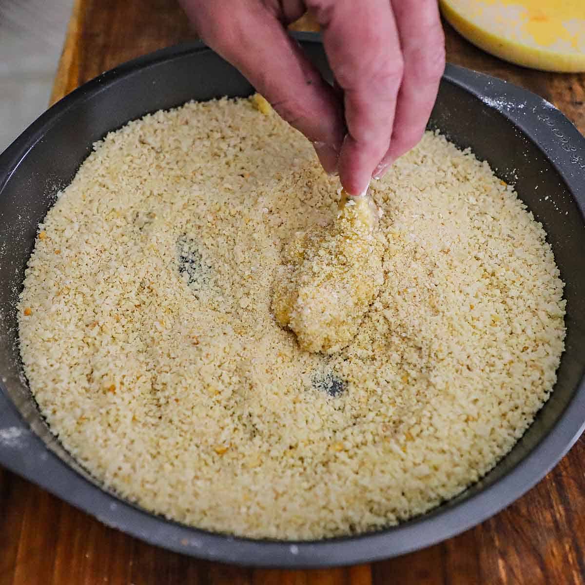A person holding a butterflied extra-large uncooked shrimp and dredging it through a breadcrumb mixture in a round baking pan.