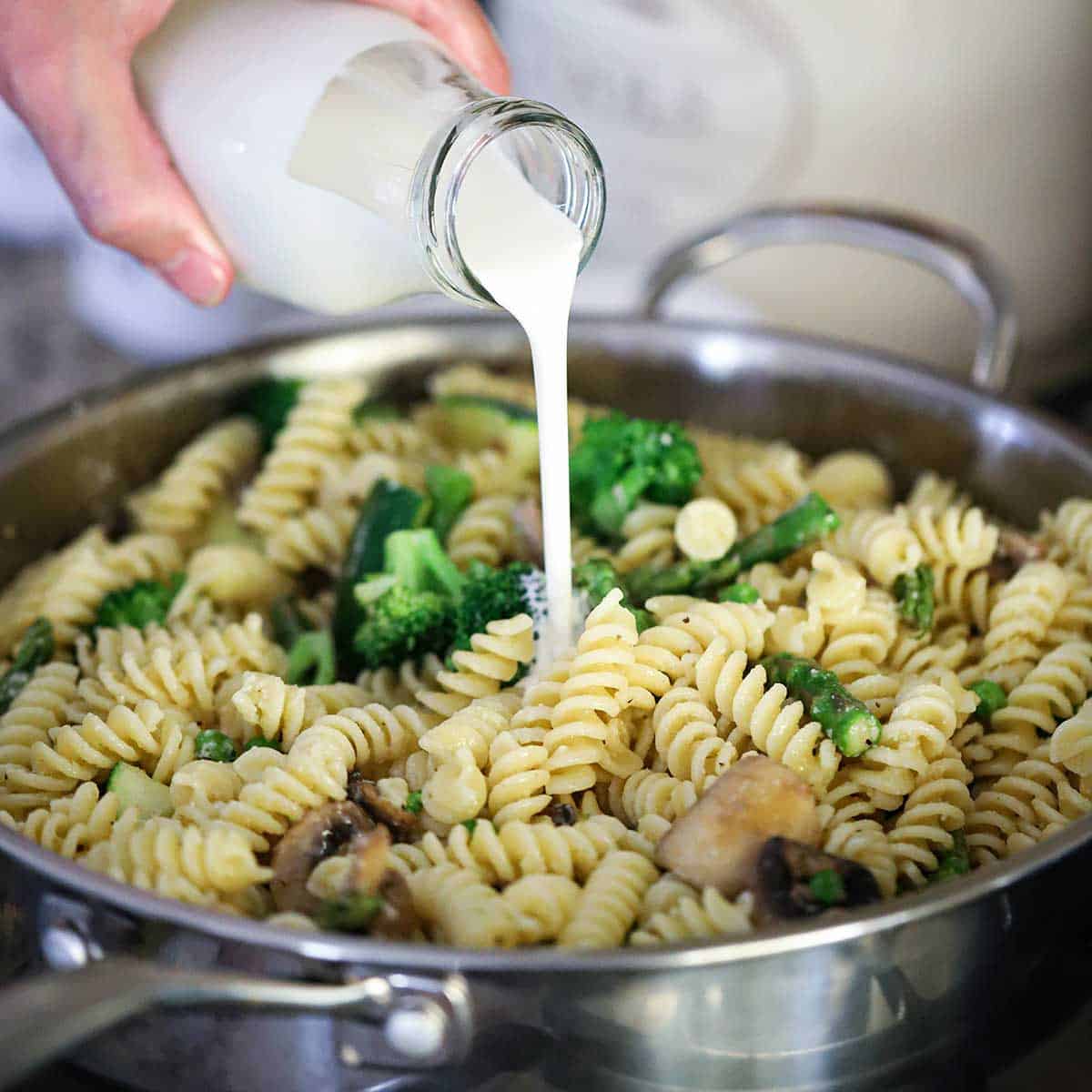 A person pouring heavy cream from a small glass milk jug into a skillet filled with cooked pasta and sautéed vegetables.