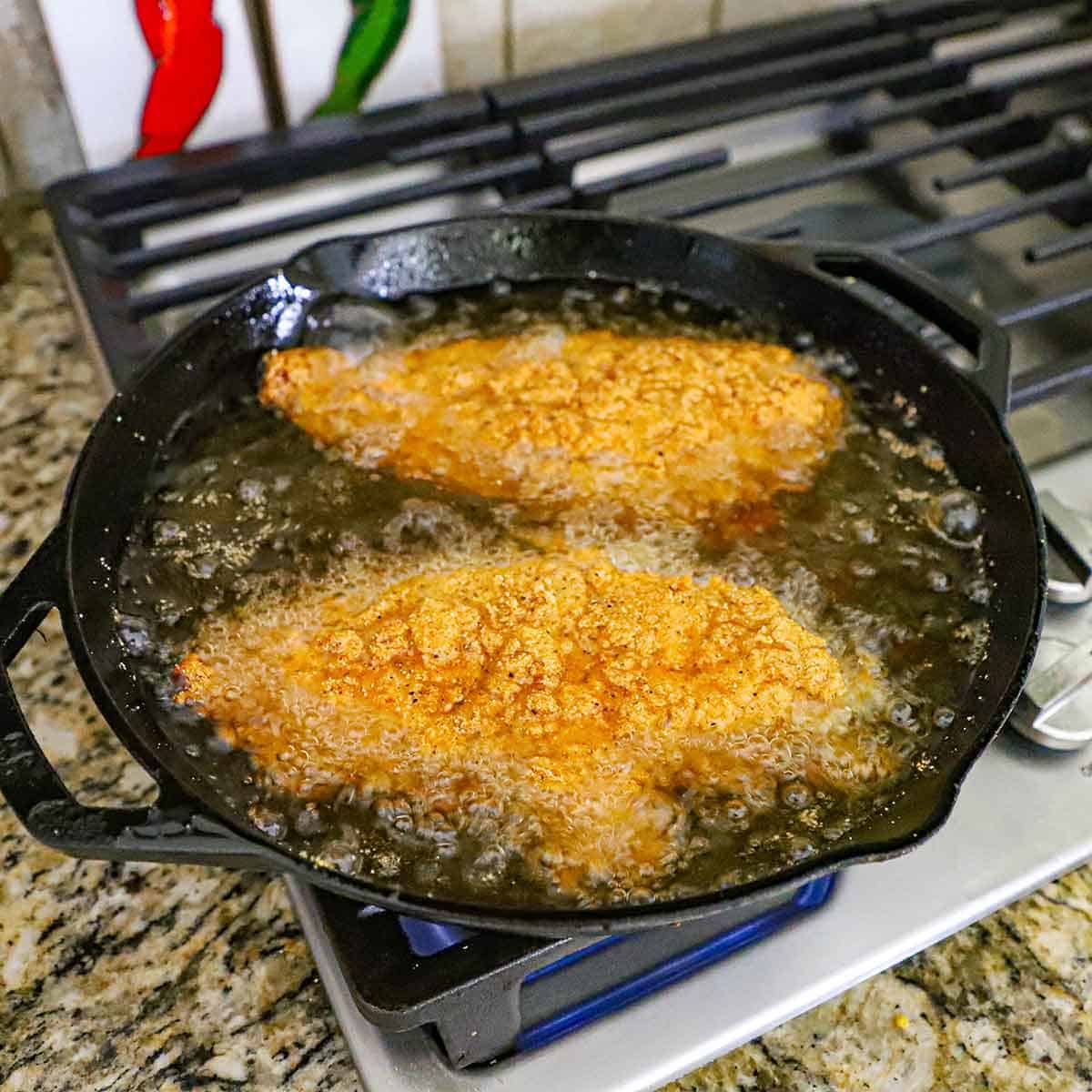 A straight-on view of two large catfish filets that are breaded and being fried in hot oil in a large cast-iron skillet on a gas stove.