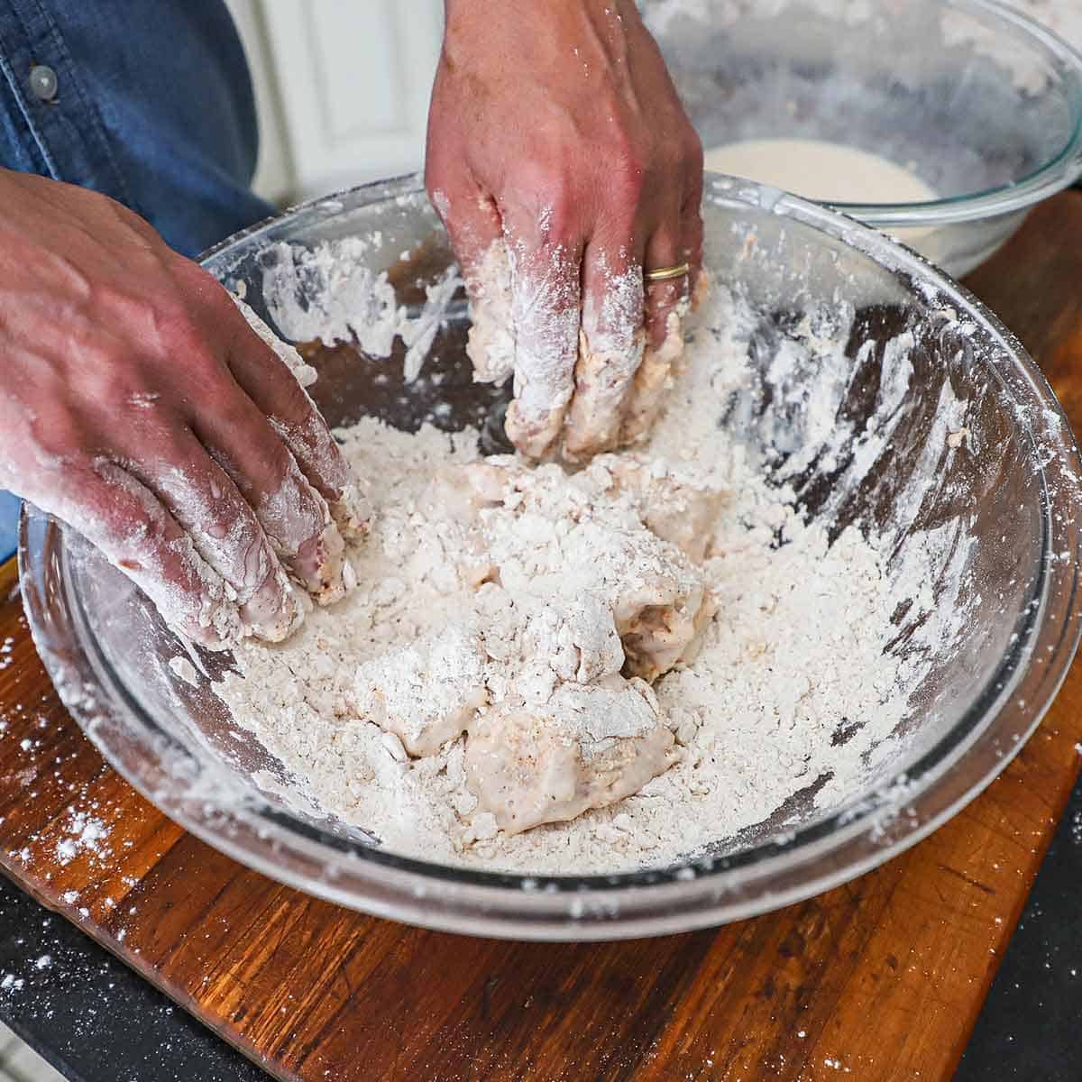 A person using his hands to toss double-coated chunks of chicken breast in a large bowl filled with seasoned flour.