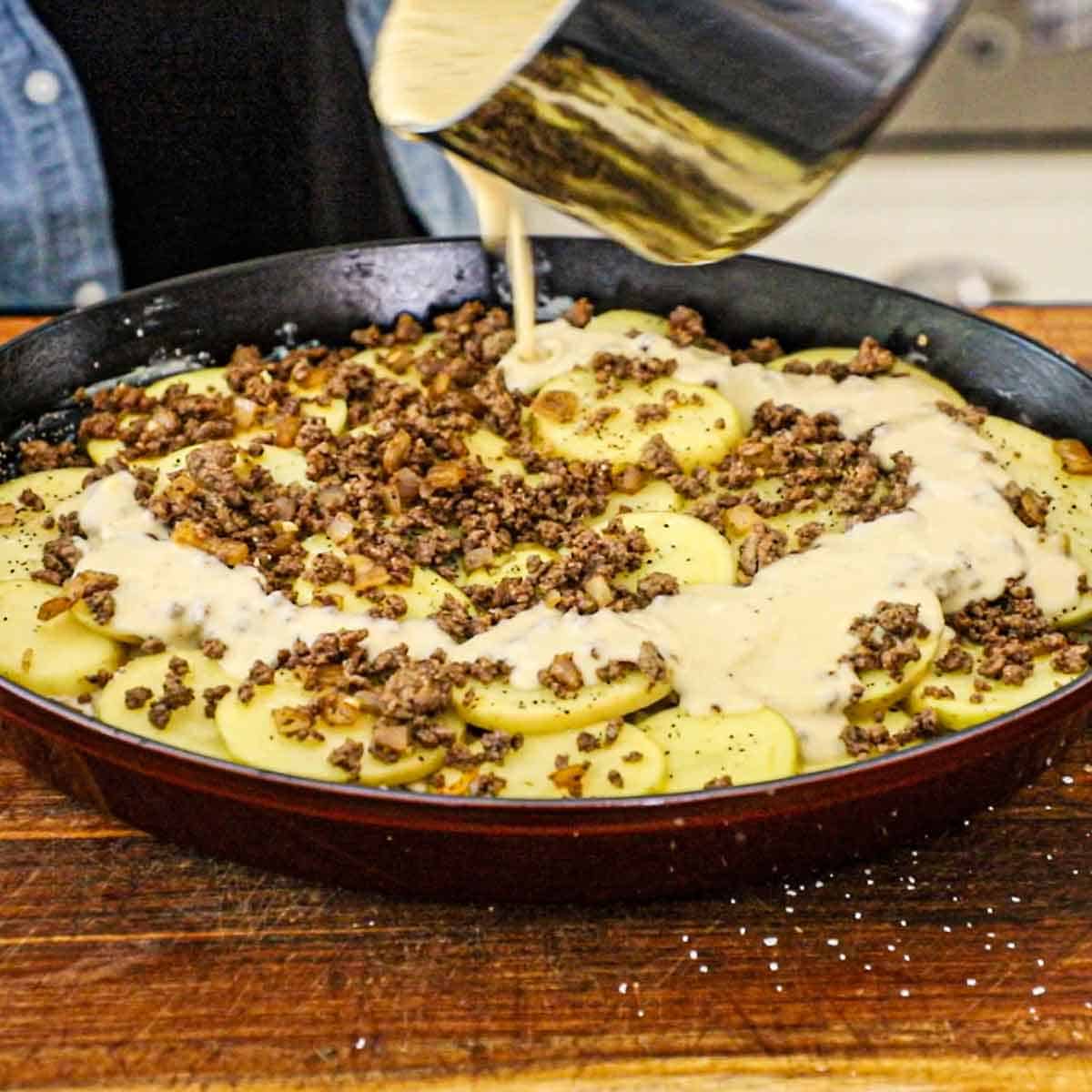 A person pouring a cheesy cream sauce from a saucepan into a large oval casserole dish that is filled with layers of sliced potatoes and seasoned and cooked ground beef.