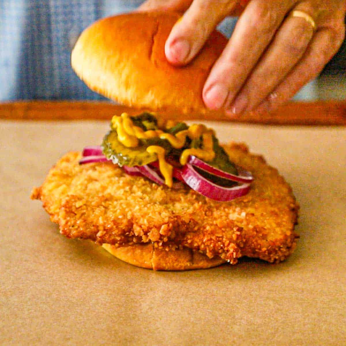 A person placing a hamburger bun on top of a large breaded pork tenderloin cutlet topped with sliced red onions, bread and butter pickles, and yellow mustard.