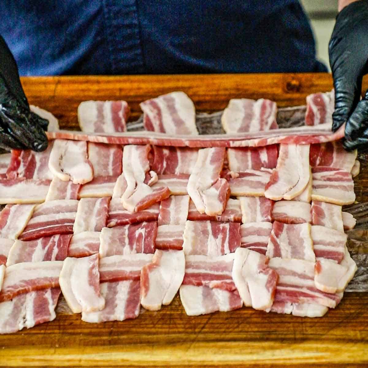 A person using his hands to create a weave of extra-thick bacon strips on a large wooden cutting board.