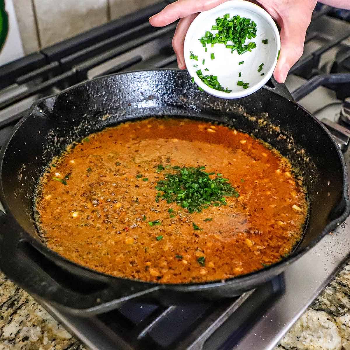 A person transferring chopped fresh herbs into a cast-iron skillet that is filled with a simmering Cowboy butter sauce.