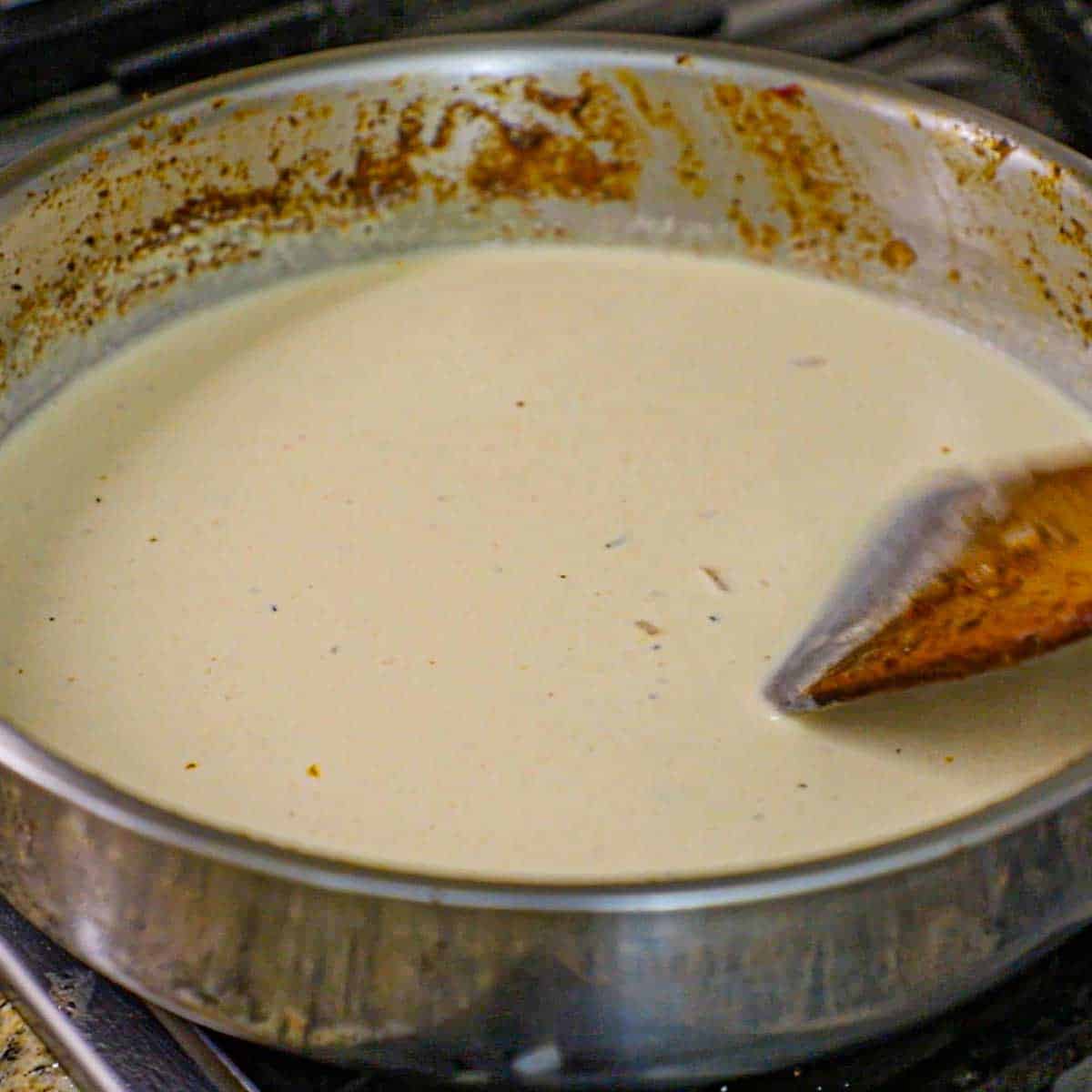 A person using a wooden spatula to stir a seasoned cream sauce in a large skillet over a gas stove.