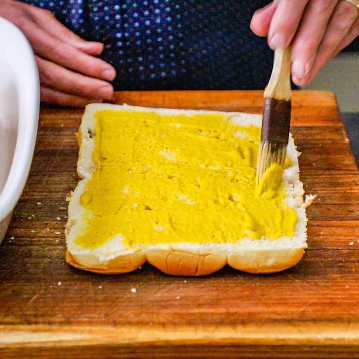 A person using a pastry brush to smear yellow mustard of the cut-side of the top portion of a rack of Hawaiian slider buns that have been split in half.