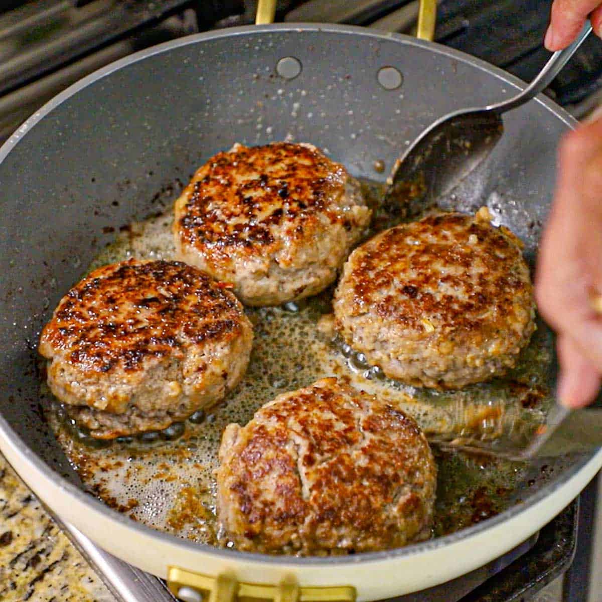 A person using a metal spatula and spoon to carefully flip over a large Dutch meatball that have been browned and seared in a skillet with melted butter on a gas stove.