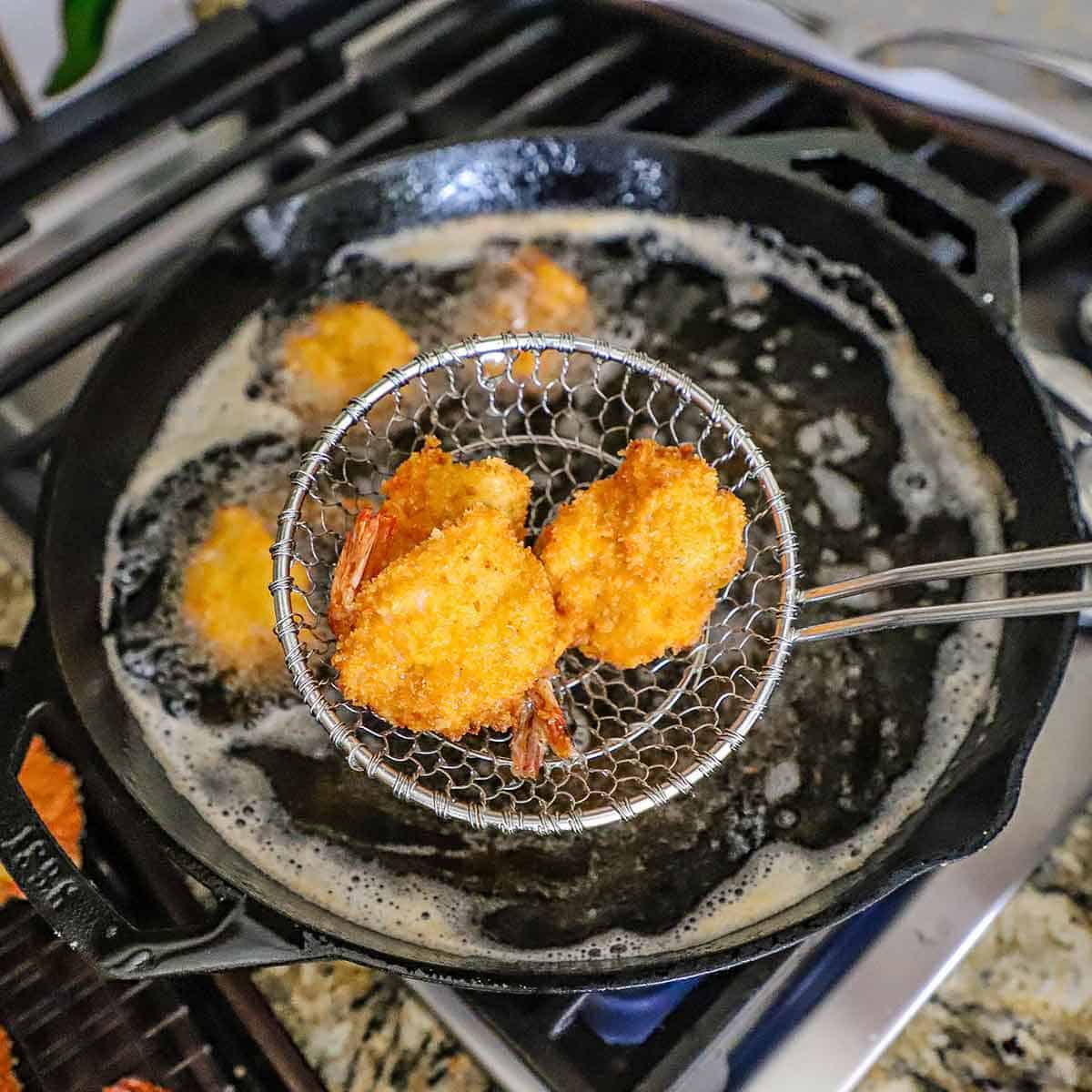 A metal spider being used to raise two fried butterfly shrimps from a large cast-iron skillet that contains hot oil and other fried shrimps on a gas stove.