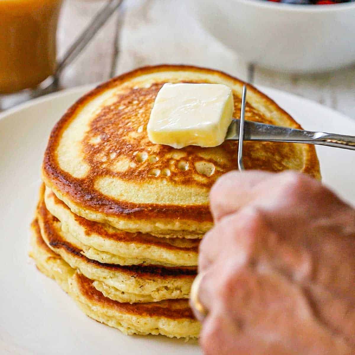 A person using two small butter knives to place a pad of butter on the top of a stack of fluffy buttermilk pancakes on a white dinner plate.