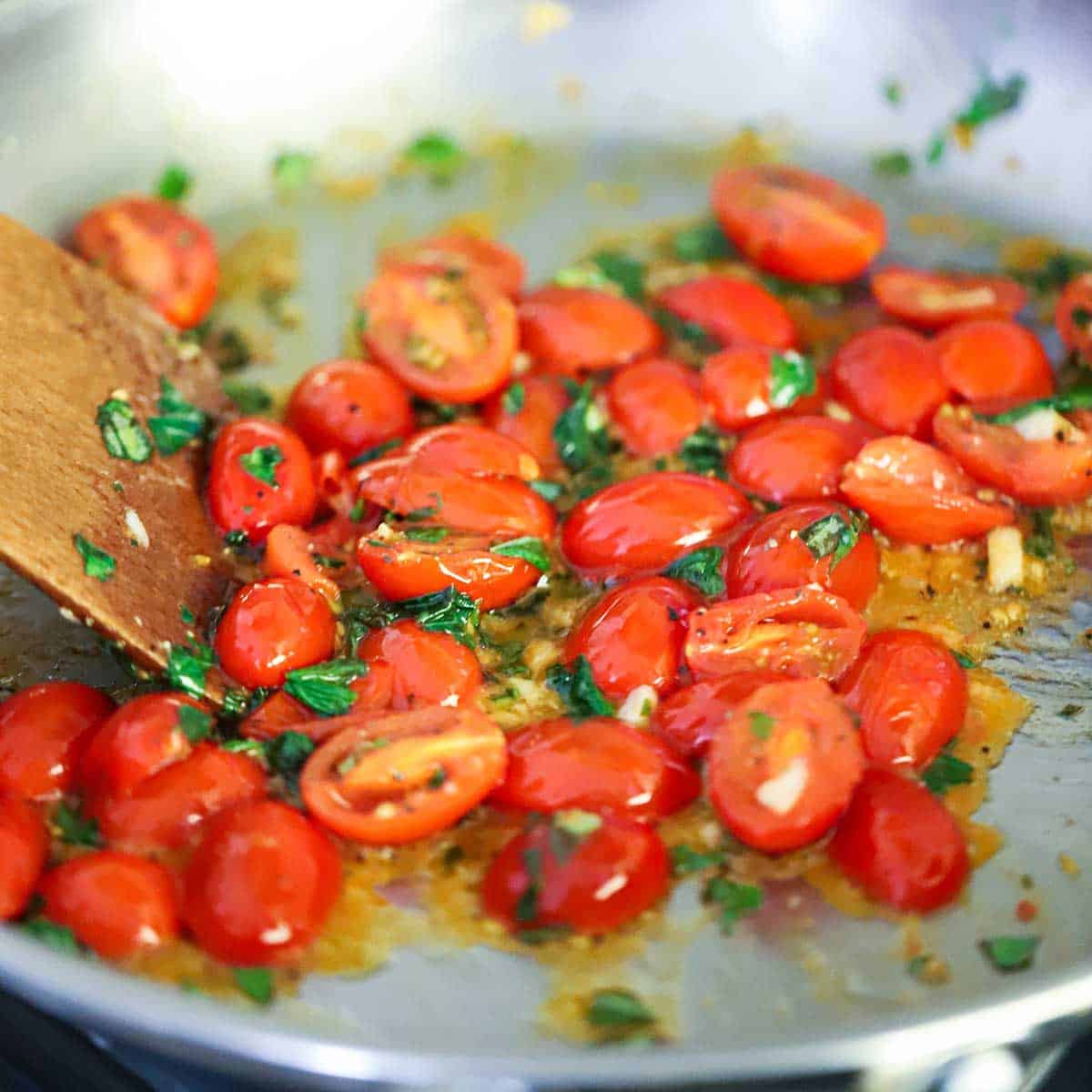A close-up view of halved cherry tomatoes that are being sautéed in a skillet along with olive oil, garlic, and fresh basil.