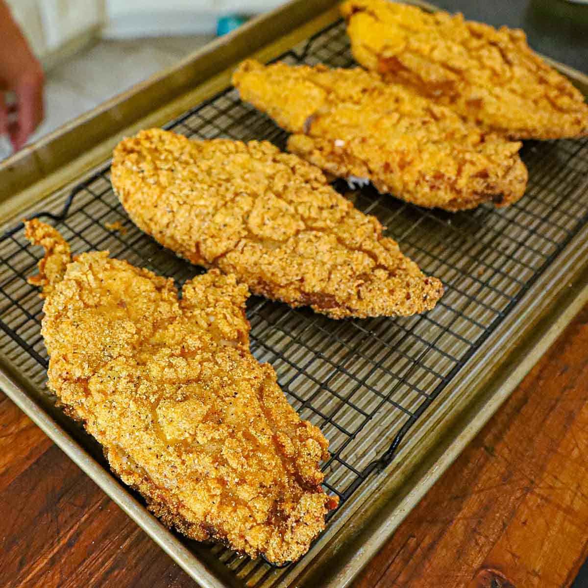 Four freshly prepared Southern fried catfish filets resting on a baking rack nestled into a baking pan on a wooden cutting board.