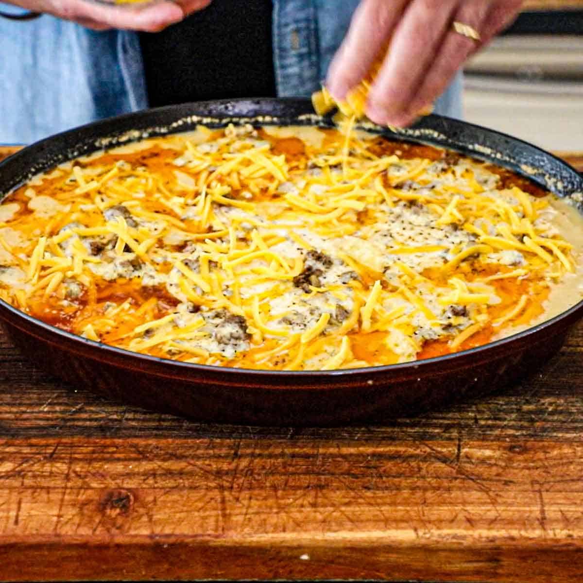 A person sprinkling shredded cheese over the top of a bubbling hamburger potato casserole that is fresh out of the oven.