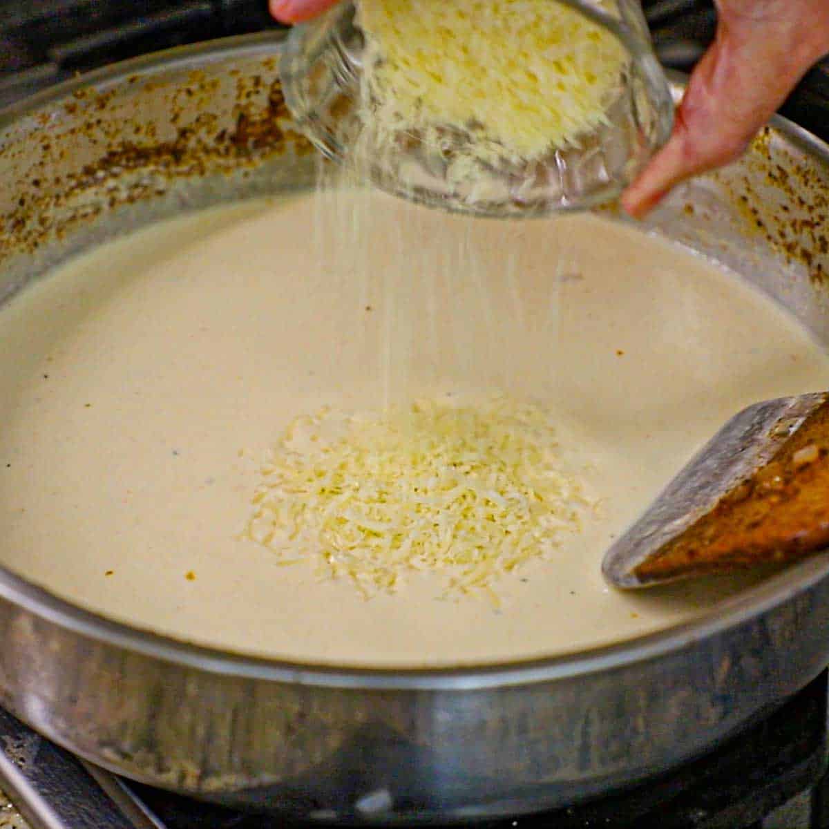 A person pouring grated Monterey Jack cheese into a large silver skillet filled with a simmering seasoned skillet.