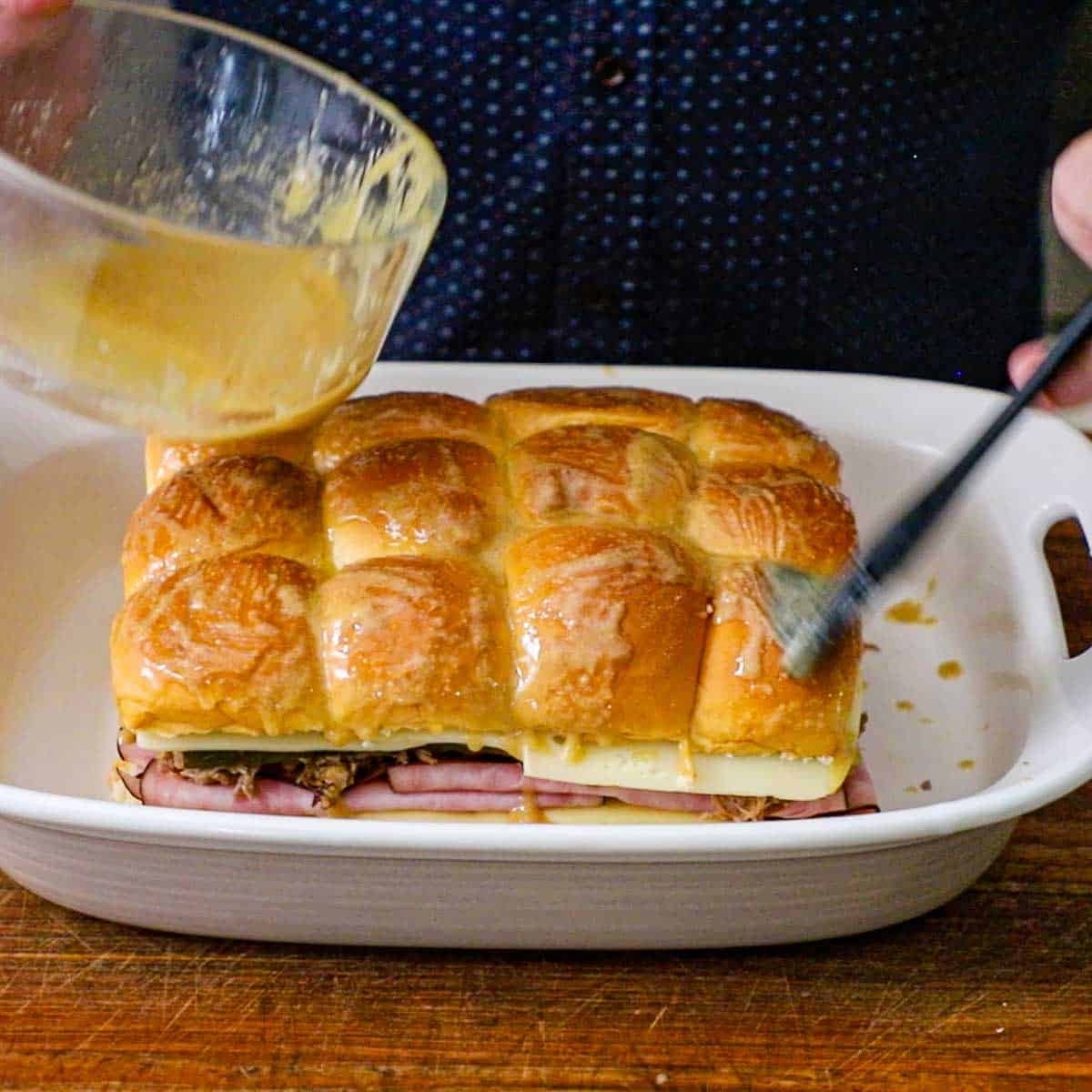 A person using a brush to apply a seasoned butter and Dijon glaze over the top of uncooked Melty Cuban Slider in a 9 by 13 baking dish.