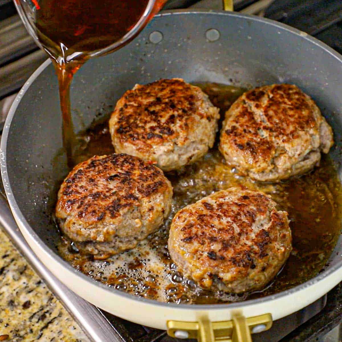 A person pouring beef stock from a glass measuring cup into a large non-stick skillet holding four large Dutch meatballs that have been seared all over on a gas stove.
