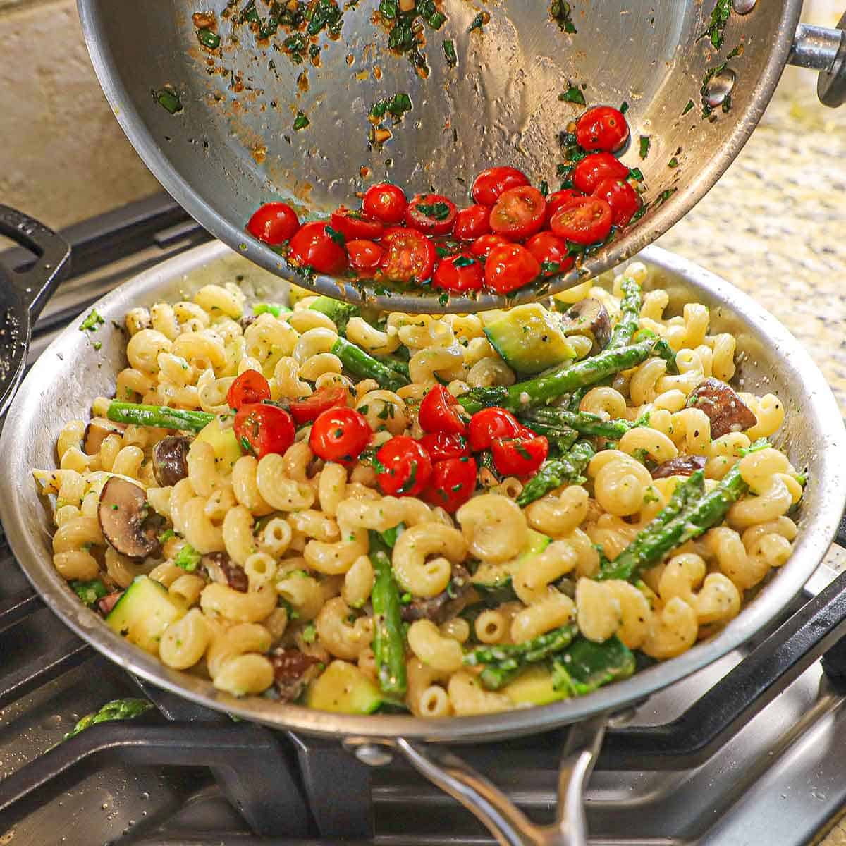 A person transferring sautéed cherry tomatoes with basil and garlic onto a skillet filled with freshly prepared pasta primavera.