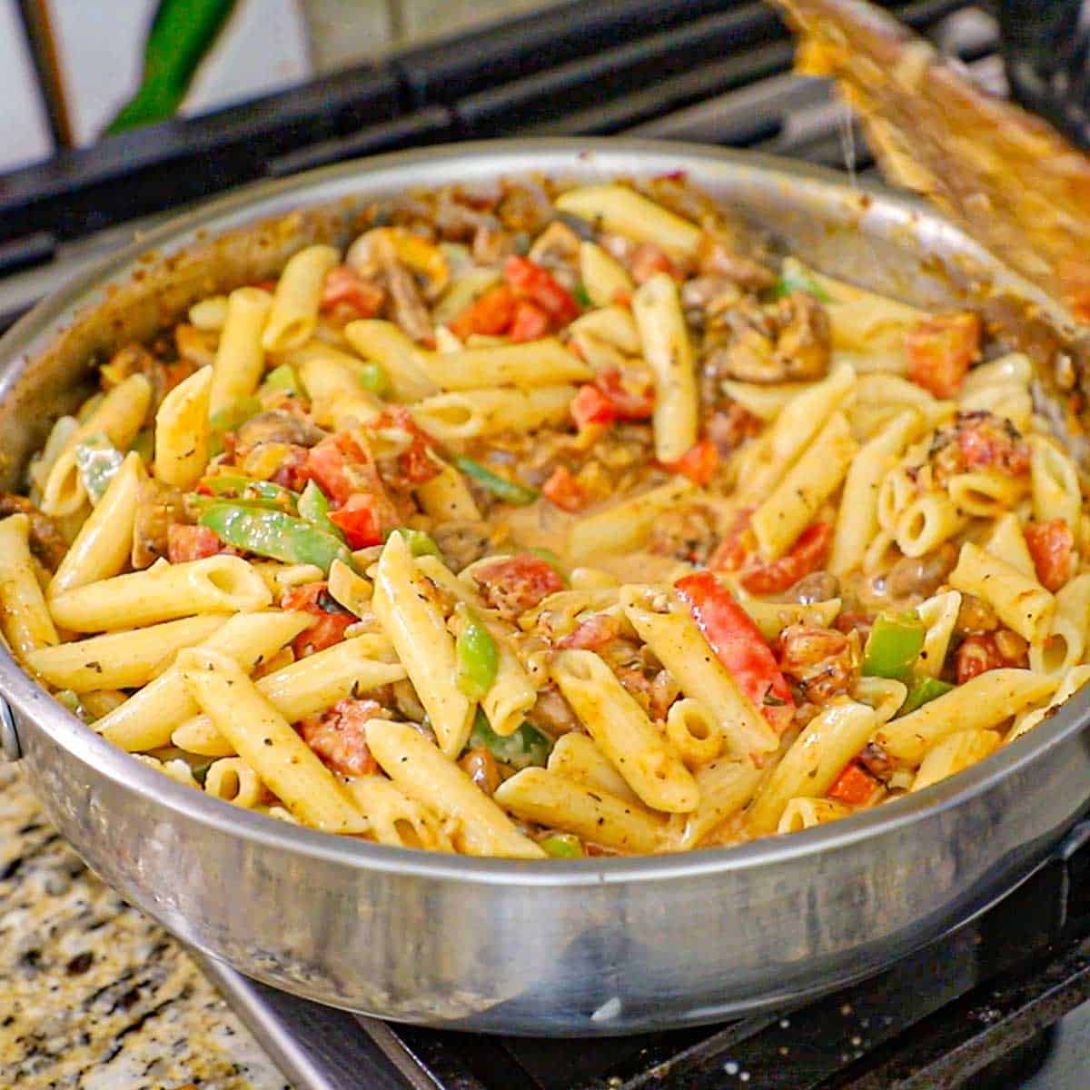 A large stainless skillet filled with a Creamy Cajun Chicken and Sausage Pasta that is being tossed with a wooden spatula.