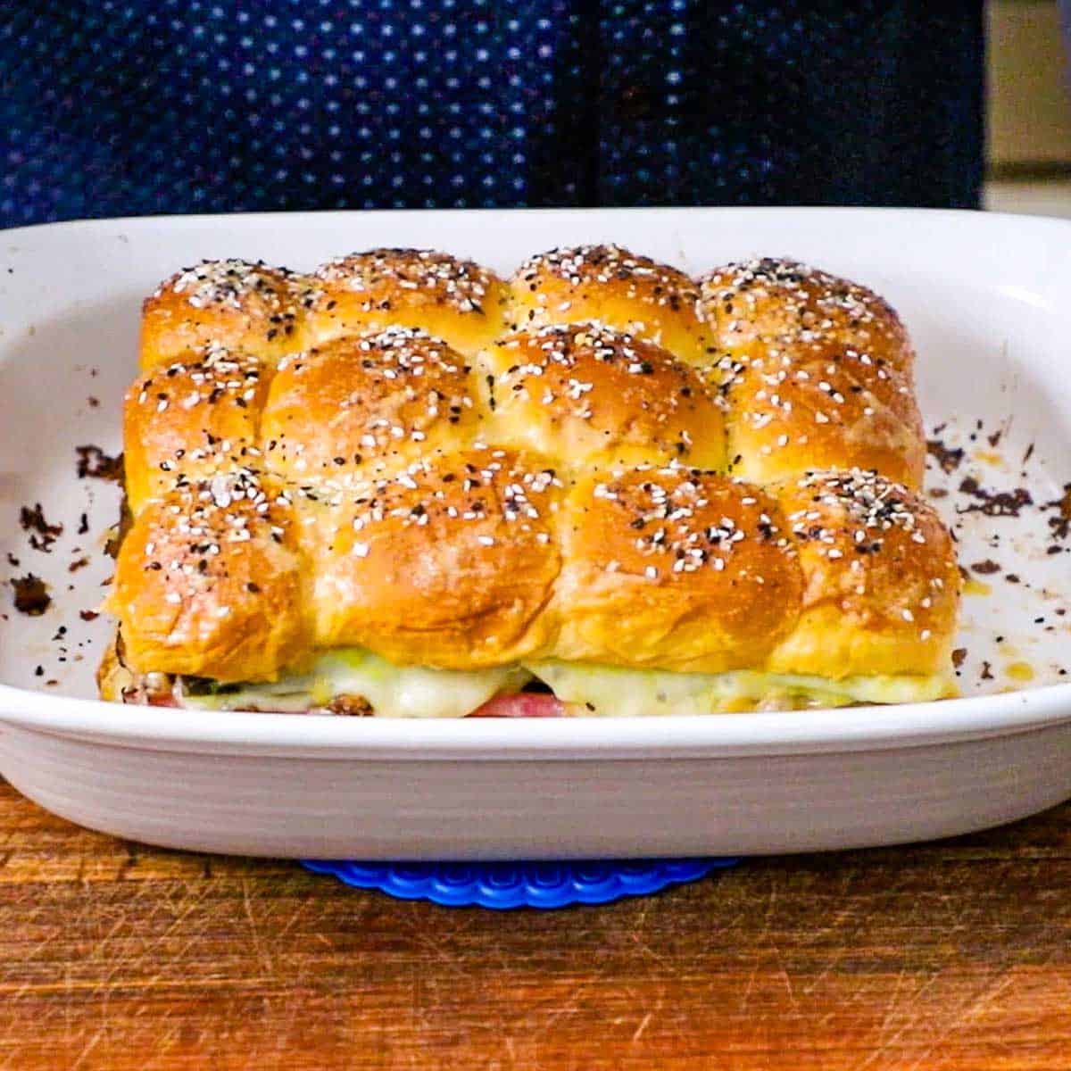A person placing a 9 by 13 baking dish containing freshly baked Cuban Sliders onto a hot pad on a wooden cutting board.