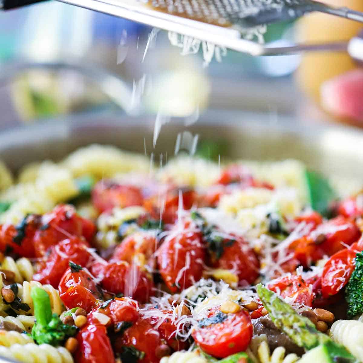 A person using a microplane to grate a block of Parmesan cheese over a skillet filled with homemade pasta primavera.