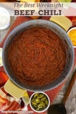 An overhead view of a large pot filled with a freshly prepared batch of Weeknight Beef Chili with a small bowl of sliced jalapeños and cornbread both nearby.