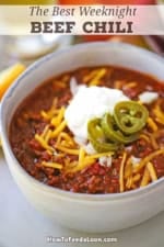 A close-up view of a white soup bowl filled with a serving of Weeknight Beef Chili garnished with shredded cheddar cheese, chopped onions, sour cream, and sliced pickled jalapeño peppers.