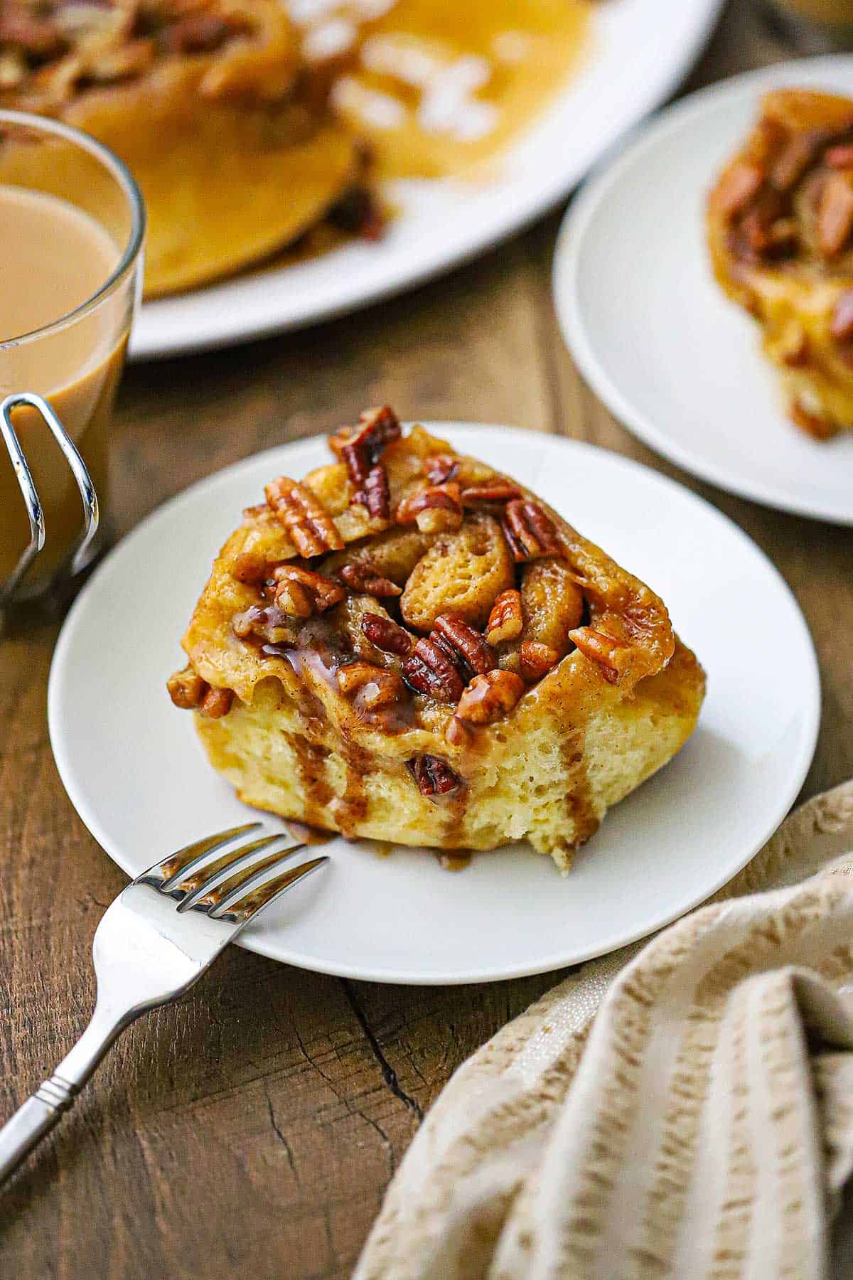 A straight-on view of a single Caramel Pecan Sticky Bun resting on a white plate with a clear glass of coffee sitting nearby.