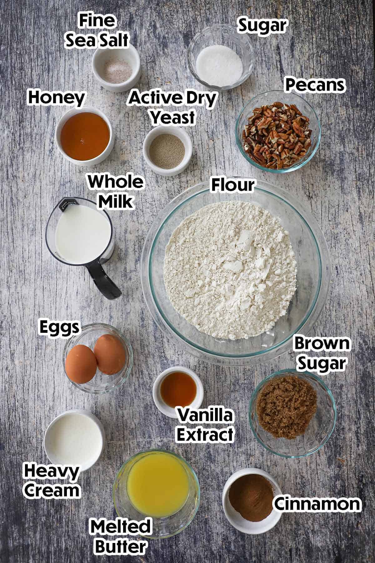An overhead view of ingredients for Caramel Pecan Cinnamon Sticky Buns on a grey wooden background including flour, milk, yeast, sugar, salt, honey, eggs, sugar, vanilla, melted butter, cream, pecans, and cinnamon.