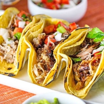 A close-up view of three double-stacked slow cooker carnitas tacos resting upright on a metal taco holder.