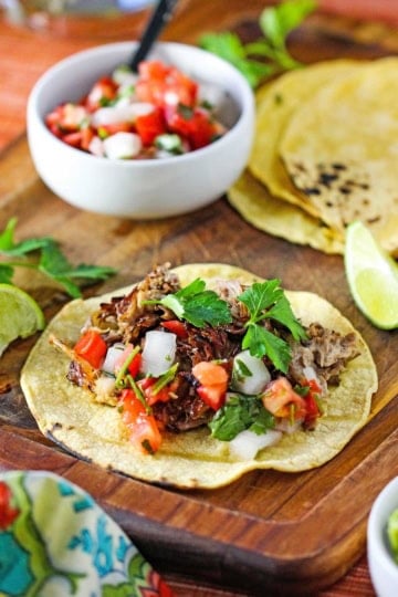 A wooden cutting board with a flat grilled corn tortilla filled with a piled of slow cooker carnitas and garnished with pico de gallo and cilantro leaves.