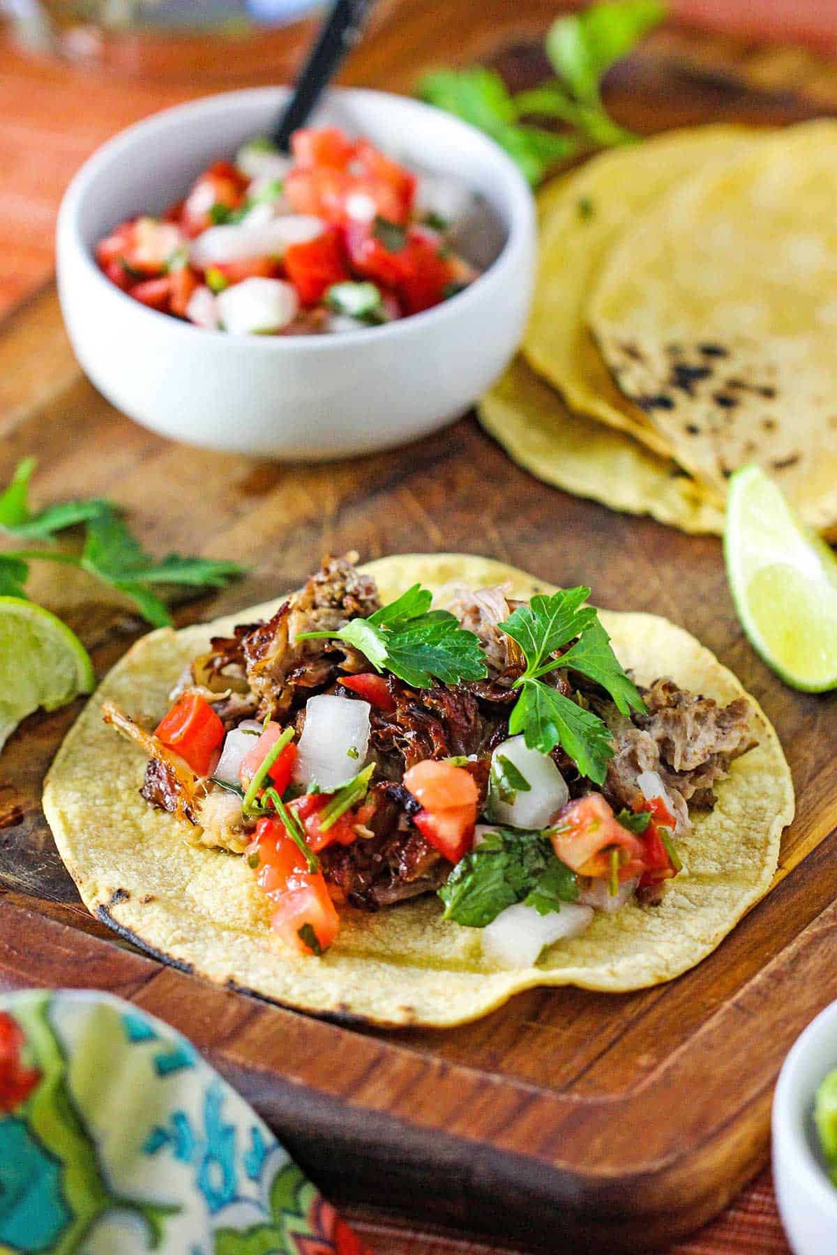 A wooden cutting board with a flat grilled corn tortilla filled with a piled of slow cooker carnitas and garnished with pico de gallo and cilantro leaves.