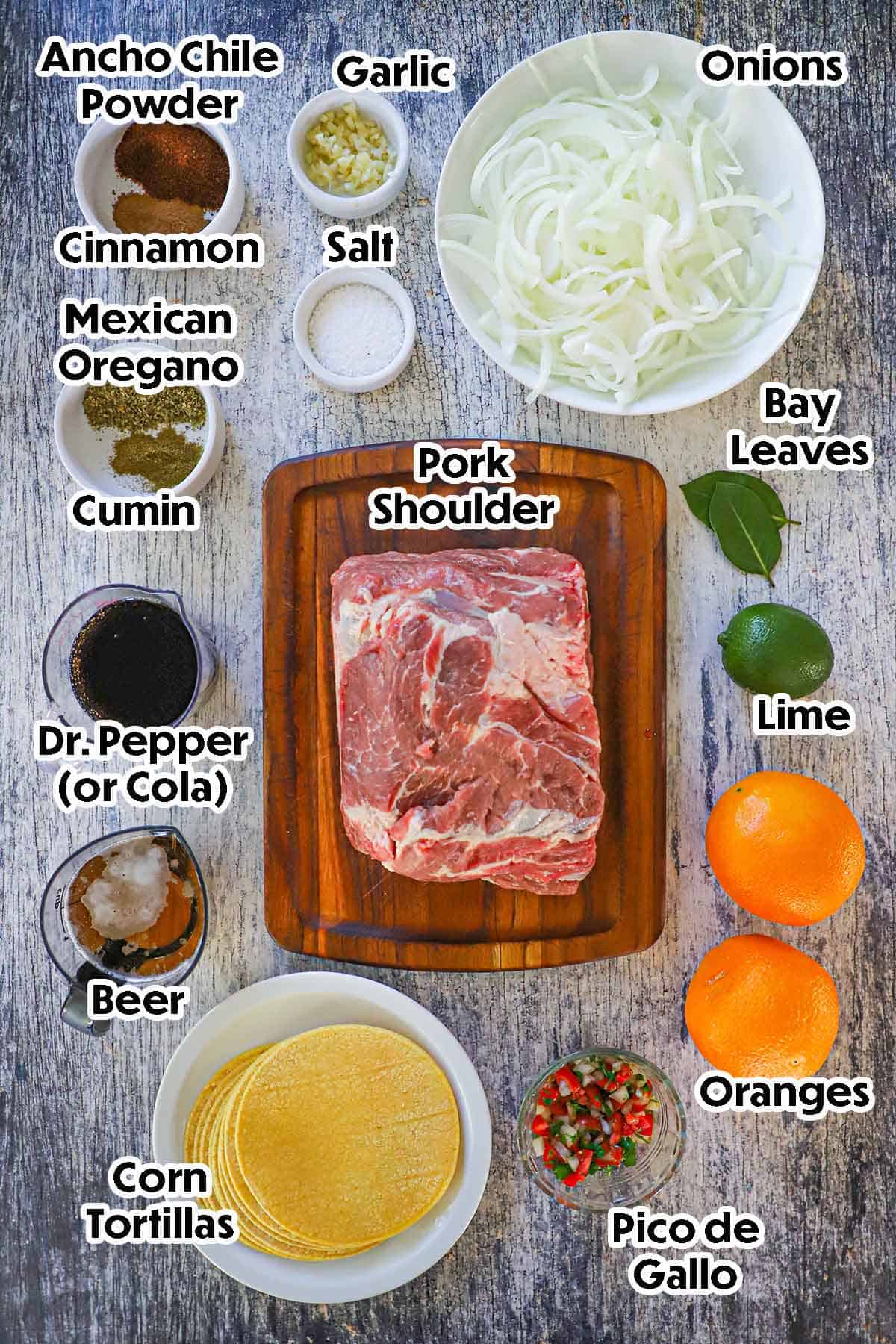 An arrangement of ingredients for slow cooker carnitas tacos on a grey wooden background including pork shoulder, onions, garlic, a lime, oranges, beer, cola, tortillas, pico de gallo, spices, and herbs.