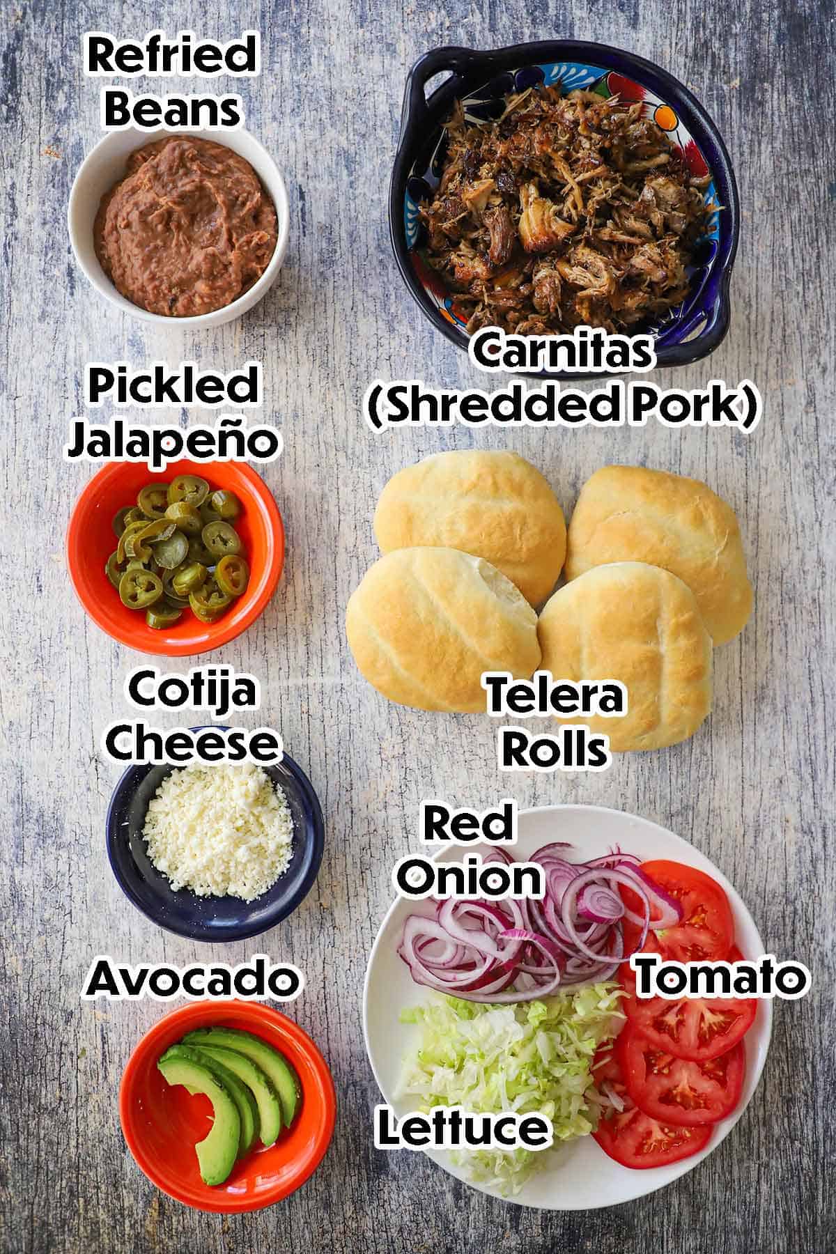 An arrangement of ingredients for carnitas torta on a grey wooden background including shredded pork, refried beans, telera rolls, jalapeños, Cotija cheese, avocado, lettuce, onion, and sliced tomatoes.