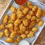 A close-up view of a metal tray lined with white wax paper and filled with freshly prepared fried cheese curds along with a small bowl of marinara in one corner and another small bowl of ranch dressing in the opposite corner.