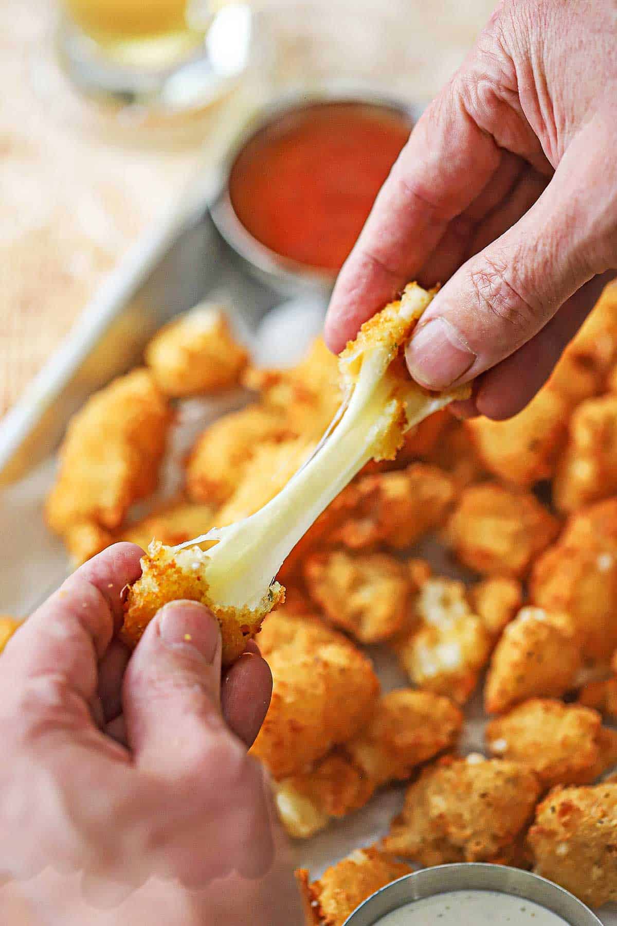 A person using two hands to pull the ends of a fried cheese curd revealing long, stretchy strand of cheese between the two crispy ends of the curd.