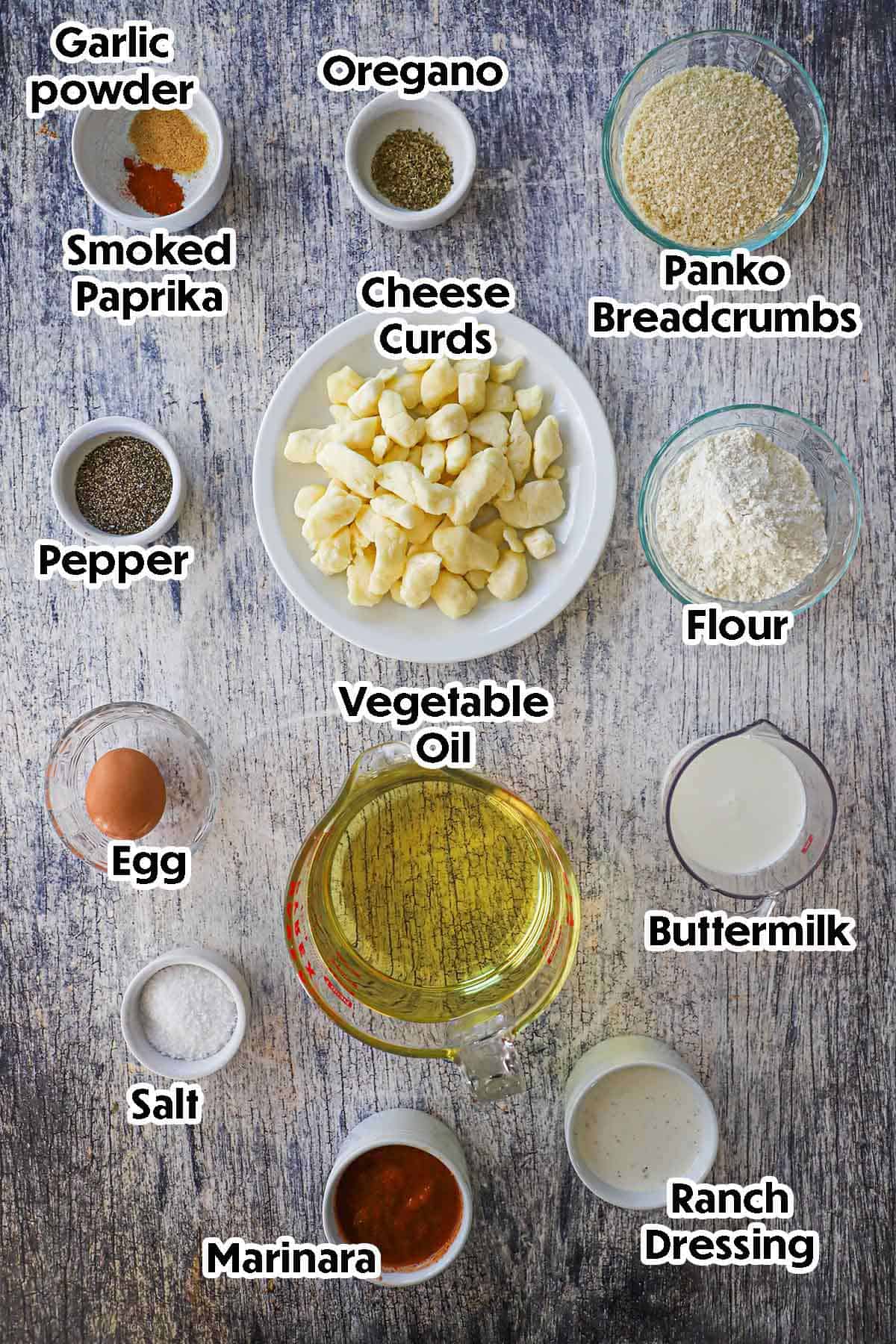 An arrangement of ingredients for Fried Cheese Curds on a grey wooden background including cheese curds, flour, breadcrumbs, buttermilk, an eggs, oil, seasonings, marinara, and ranch dressing.