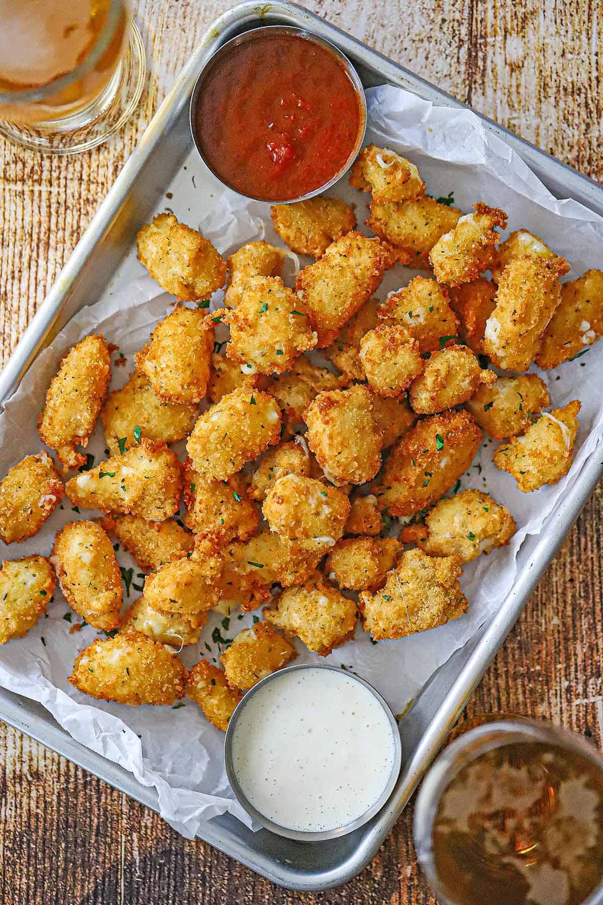 An overhead view of a metal tray lined with white wax paper and filled with freshly prepared fried cheese curds along with a small bowl of marinara in one corner and another small bowl of ranch dressing in the opposite corner.