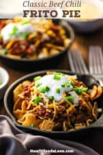 A close-up, straight-on view of two small cast-iron skillets both filled with individual servings of Classic Frito Pie topped with Beef Chili, shredded cheese, sour cream, and chopped jalapeños.