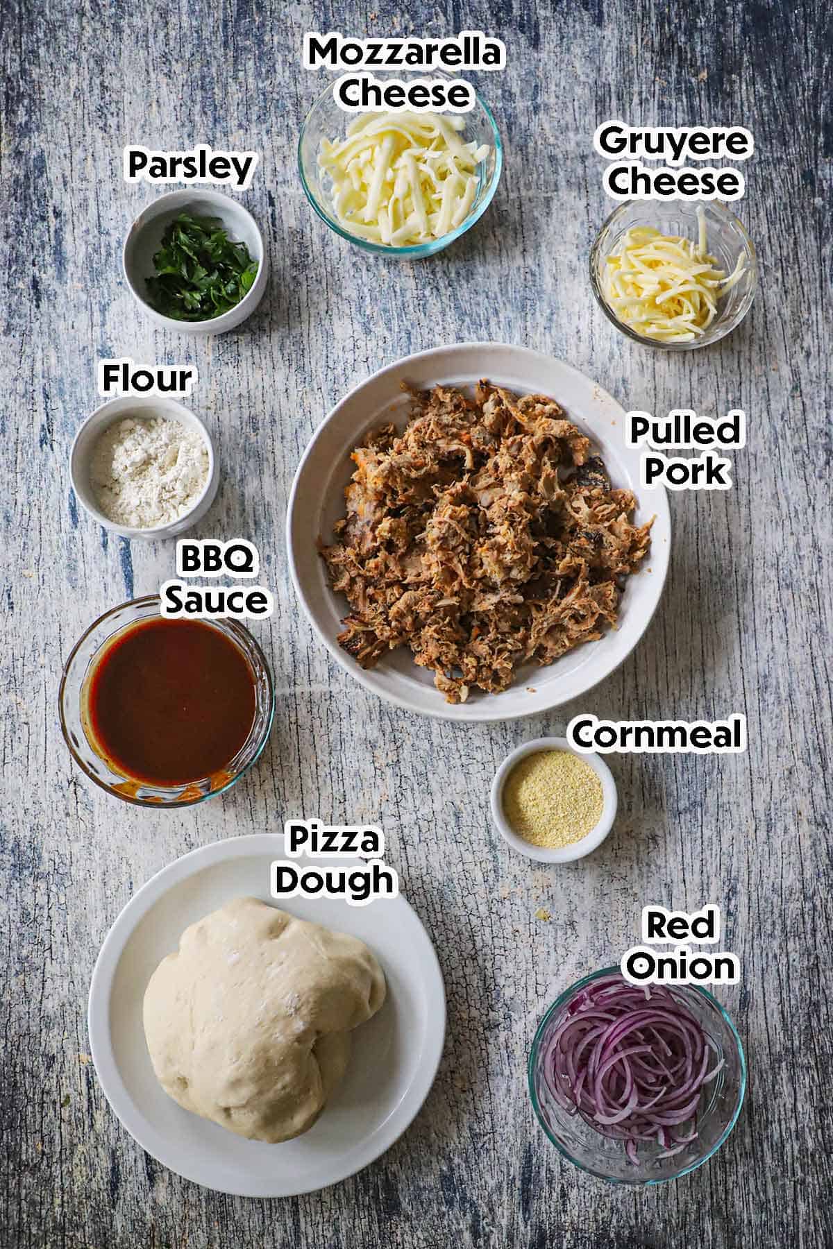 An arrangement of ingredients for Grilled Pulled Pork Pizza on a grey-wooden background including pulled pork, pizza dough, BBQ sauce, onion, flour, Mozzarella cheese, gruyere cheese, and cornmeal.
