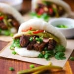 A close-up view of a Braised Pork Belly Bao Buns each resting on a square piece of brown paper on a large wooden cutting board.