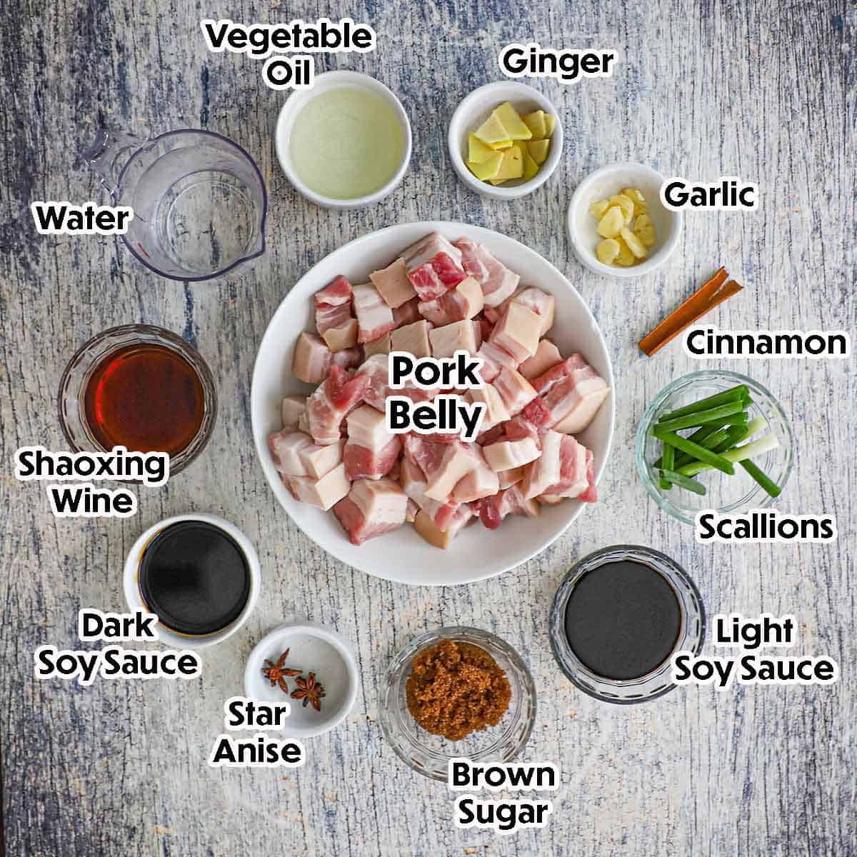 An arrangement of ingredients for Asian Braised Pork Belly on a grey wooden background including chunks of pork belly, ginger, garlic, cinammon, scallions, oil, water, Shaoxing wine, spices, and seasonings.