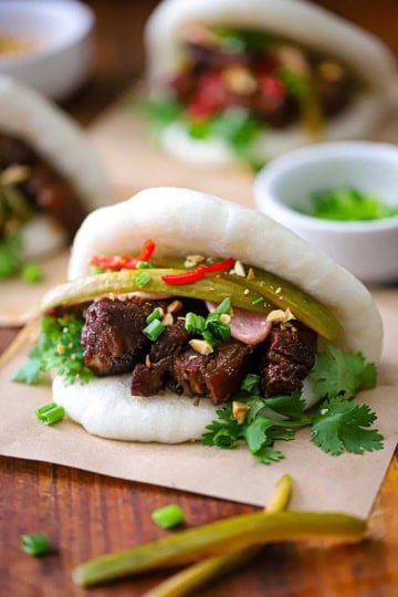A straight-on view of aBraised Pork Belly Bao Buns each resting on a square piece of brown paper on a large wooden cutting board.